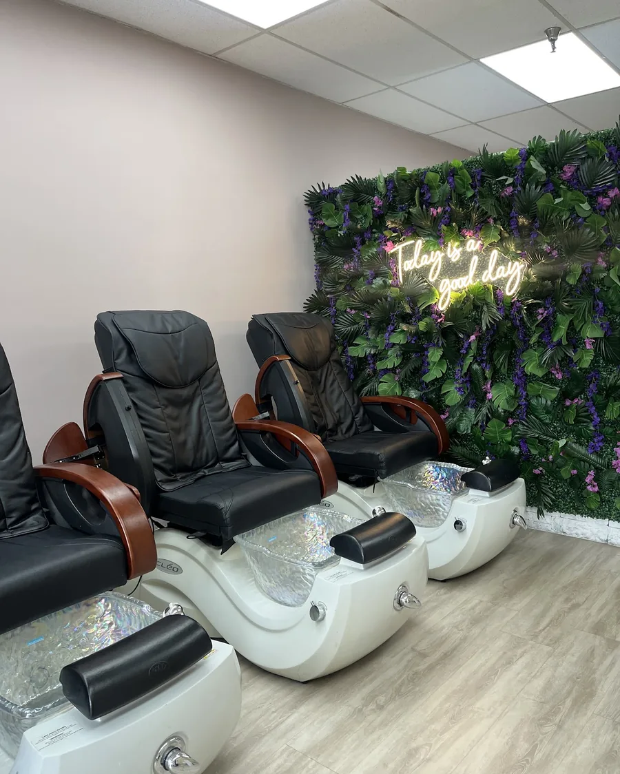 Three black leather massage pedicure chairs in front of a green foliage wall with a warm neon sign