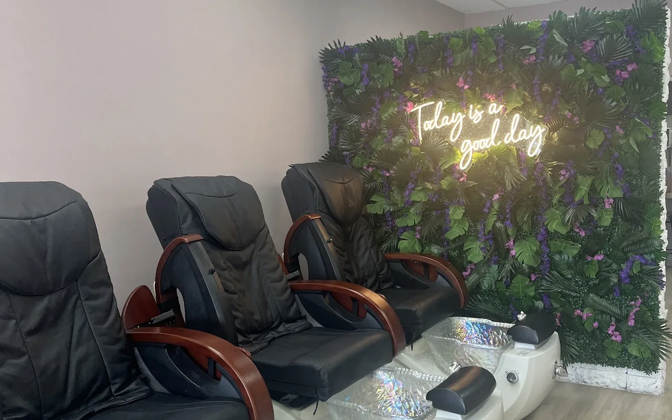 The pedicure area at Victoria's Nails and Spa: black massage chairs facing a wall of green foliage lit by a warm neon script sign